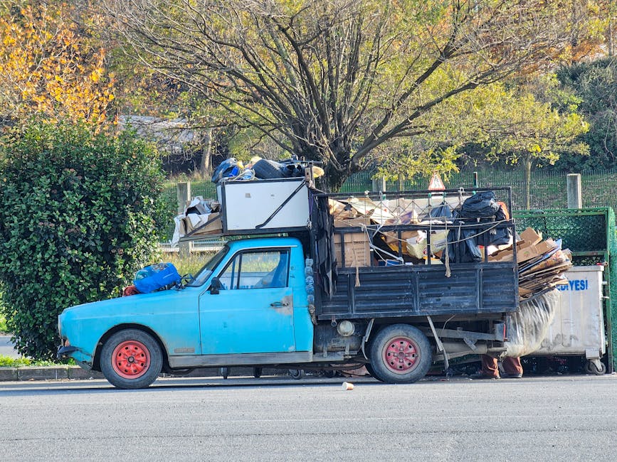 A small, vintage blue flatbed truck parked parallel to a curb in an outdoor environment. The truck's cab is painted in a faded, light blue color, with visible signs of rust and wear on the door and front fender. The flatbed section is constructed with black metal panels and is heavily loaded with a variety of household waste, including large cardboard boxes, black plastic trash bags, and miscellaneous debris, some of which is loosely tied with ropes. A white refrigerator or appliance is positioned towards the front of the load, partially obscured by other waste. The truck's red painted steel wheels are visible, with the rear wheel partially supporting the weight of the piled debris. Behind the truck, a lush green tree with budding leaves provides a natural backdrop, indicative of late autumn or early spring. To the left, a well-maintained hedge separates the roadside from a garden or park area, while in the background, a metal fence and additional trees contribute to the outdoor setting. The scene is evenly lit with natural daylight, suggesting clear weather. The image relates to the context of alternative waste collection or private rubbish removal services, reflecting a typical scenario where a small-scale rubbish removal company such as Rubbish Removal Bromley handles uncollected household waste and bulky items outside local authority collection areas.