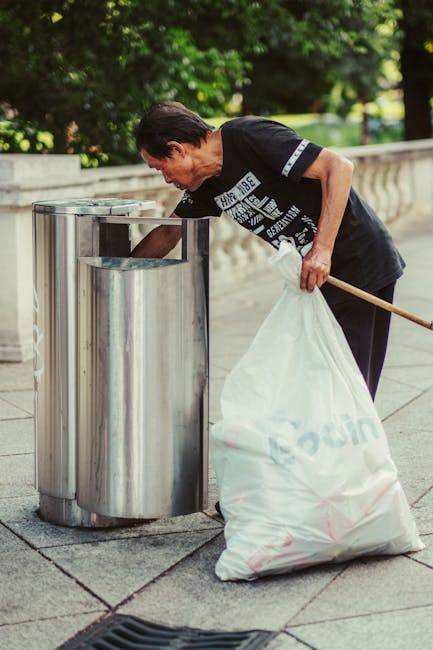 A middle-aged man with dark hair and light skin is seen bending over a cylindrical, stainless steel rubbish bin on a paved pathway in an outdoor setting. He is wearing a black T-shirt with light-colored text and graphics on the front, along with dark trousers. The man is holding a large, white plastic rubbish bag filled with waste, which is partially open and resting on the ground, while his other hand is reaching into the bin. A wooden-handled litter picker or grabber is tucked under his arm, suggesting he is engaged in waste collection or rubbish removal. In the background, there are lush green trees and foliage, indicating that the location is a park or garden area. The lighting is natural daylight, casting soft shadows on the scene, and the atmosphere appears calm and organized, reflecting a community effort or private waste handling activity aligned with rubbish removal services. The overall scene exemplifies proactive waste disposal through independent collection in an outdoor environment.