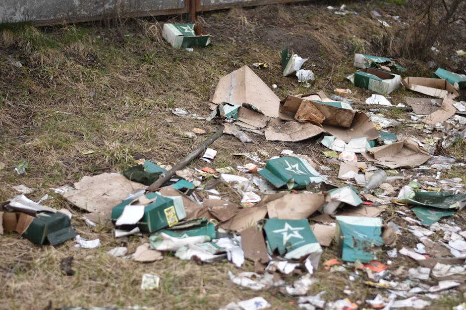 A patch of ground outdoors covered with scattered debris and rubbish, including flattened cardboard boxes in green and brown with visible creases and torn edges, some displaying a white star logo. The boxes are partially crushed and layered among crumpled paper and small plastic waste. The surrounding area has short grass and patches of bare soil, with a wooden fence at the top left corner. The debris is spread across a slight incline, with some pieces near the base of the fence and others further down, creating a cluttered scene relevant to waste disposal and rubbish clearance services. Natural light illuminates the scene, highlighting the textures of the shredded cardboard and various waste materials, indicative of an unsightly area awaiting professional rubbish removal, such as that provided by Rubbish Removal Bromley.