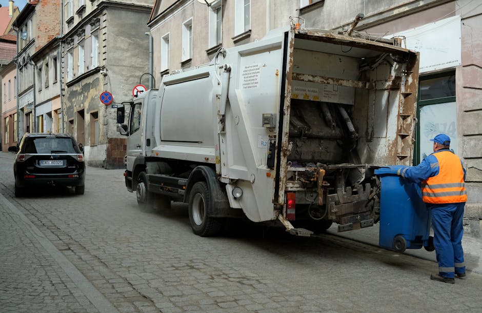 A cluttered collection of overflowing rubbish bins and scattered household waste occupies a section of a paved parking area in front of a commercial building. The waste includes various types of refuse such as black plastic garbage bags, flattened cardboard boxes, crumpled paper, and enclosed cartons. The grey and black bins are filled to capacity, with some waste spilling over and lying on the ground. The grey bin, designated for mixed paper and card, and other bins with black lids are visibly surrounded by loose rubbish, including packaging materials and discarded items. In the background, a parked silver hatchback car with a partially visible license plate is positioned behind a metal barrier. The building behind features a blue scaffolding structure with a façade that includes shopfront signage with icons indicating no smoking and other warnings. The scene is illuminated by natural daylight, with the environmental context suggesting this is an area where waste collection or disposal services might be needed, highlighting the importance of proper rubbish removal possibly handled by companies like Rubbish Removal Bromley for local waste clearance.