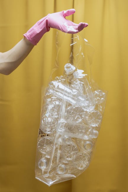 A person's hand wearing a pink glove is holding a clear plastic bag filled with crushed empty plastic bottles and containers, with some flattened. The bag is being lifted against a yellow background, with the bottles visible through the thin, transparent plastic. The contents consist mainly of transparent, glossy plastic with some ridged and smooth surfaces, typical of soft drink bottles and similar packaging. The scene depicts a waste collection or recycling activity, possibly related to rubbish removal services in Bromley, with the focus on the plastic waste being prepared for disposal or recycling. The vivid yellow backdrop provides a neutral contrast to the clear plastic and the pink glove, emphasizing the handling of household or commercial plastic waste.