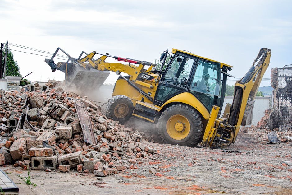 A yellow backhoe loader with a glass-enclosed cabin and large black rubber tires is actively engaged in debris removal at a construction site in an urban area. The machine's hydraulic arm extends forward, equipped with a bucket filled with broken bricks, concrete chunks, and wooden debris, which it is lifting from a pile spread across a paved surface. Dust is visible rising from the debris, indicating recent disturbance. In the background, partially demolished brick and concrete structures are visible, along with a cloudy sky overhead. The scene is characteristic of a building demolition or renovation project, and the presence of the machine and debris suggests that Rubbish Removal Bromley specializes in clearing builders waste in Bromley or nearby areas.