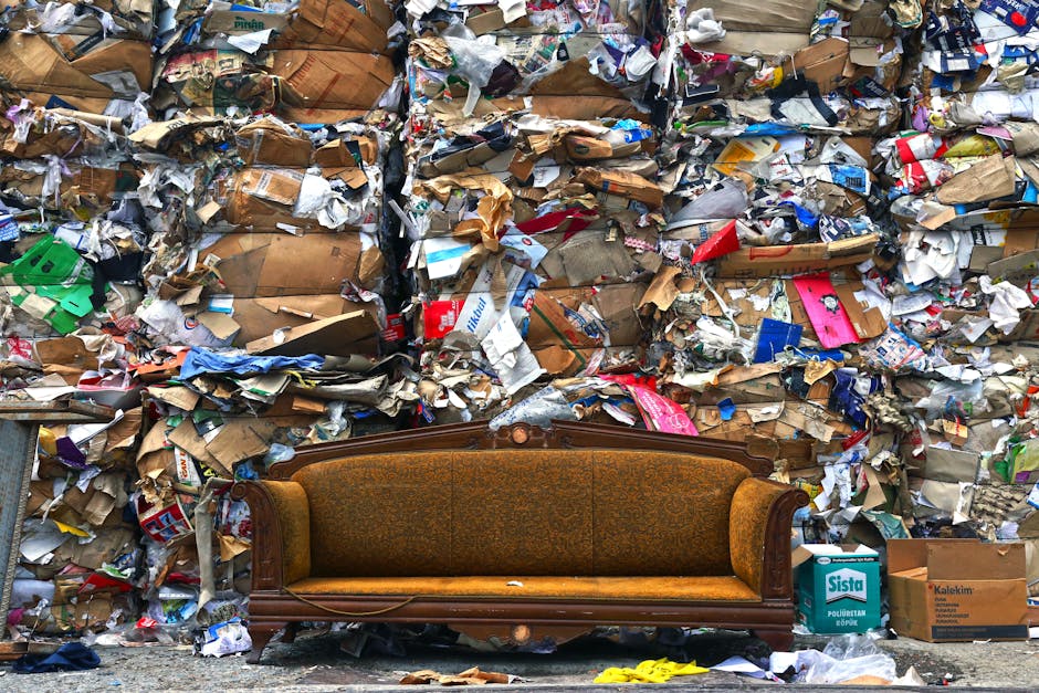 A vintage wooden sofa with upholstered armrests and a fabric seat cushion, positioned outdoors on a concrete surface, is placed in front of a large wall of tightly packed rubbish and paper waste. The wall contains layers of flattened cardboard boxes, crumpled paper, and packaging materials in various shades of brown, white, and coloured printed designs. The sofa has a dark polished wood frame with carved detailing along the top and sides, and its fabric upholstery features a muted, textured pattern in an earthy tone. Surrounding the sofa are scattered cardboard boxes, including a branded cardboard box visible on the right. The scene suggests a waste collection or disposal site typical of rubbish removal services in Bromley, with the background waste emphasizing the importance of professional rubbish clearance and furniture disposal specialists. The image is well-lit with natural daylight, highlighting the textures of the materials and the contrast between the furniture and waste environment.