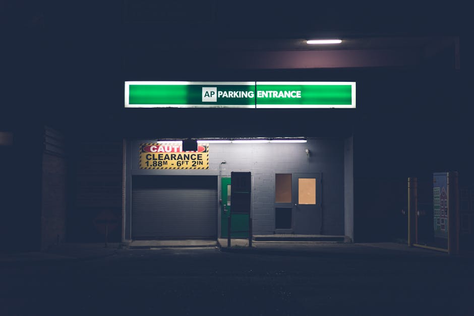 A dimly lit underground parking entrance with a large green and white illuminated sign displaying 'AP Parking Entrance' above the doorway. Below the sign, there is a yellow clearance warning sign indicating a maximum height of 6 feet 2 inches, positioned next to a grey metal roller shutter gate that is partially closed. The entrance area features a white painted brick wall, with two small windows or access panels to the right of the shutter, one covered with a brown board and the other possibly open or boarded up. To the right of the structure, there is a vending machine with a multicoloured front panel, standing on a dark paved surface. The overall scene is dark, suggesting the photo was taken at night or in low light conditions, with minimal ambient lighting penetrating the scene. The setting likely relates to a commercial or residential underground parking facility, possibly associated with a Bromley location, where rubbish removal services could involve clearing debris from such service entrances or surrounding areas.
