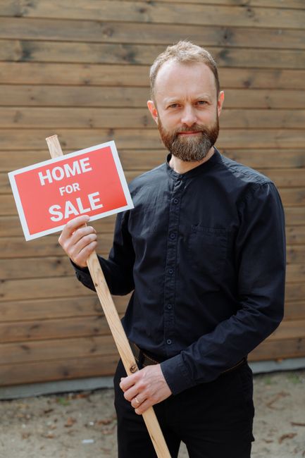 A man with a neatly trimmed beard and short light brown hair is standing outdoors against a background of horizontal wooden fencing. He is wearing a dark, long-sleeved, button-up shirt and has a neutral expression on his face. He is holding a wooden signpost with a rectangular sign attached to it, which displays a red and white sign that reads 'HOME FOR SALE' in bold letters. The sign is positioned at chest height and slightly angled towards the camera. The ground beneath him appears to be a mix of paving and dirt. The image suggests a real estate context, but it is relevant to rubbish removal services in Bromley by illustrating the type of property clearance or associated property-related activities undertaken by Rubbish Removal Bromley, especially in local residential areas near Bromley. The overall scene is brightly lit with natural daylight, and the wooden fence provides a rustic backdrop, which subtly aligns with the suburban environment of the area.