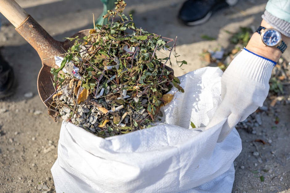 A person wearing a grey sweater, white gloves, and a wristwatch is using a small shovel to load a white woven polypropylene sack with a mixture of garden waste, including green plant clippings, small branches, and dried leaves, on a gravel surface. The sack appears partially filled, with the waste visibly protruding from the top. The background shows an outdoor area with dirt and scattered debris, typical of garden or driveway clearance work. The scene suggests a gardening or waste removal activity, possibly organized by Rubbish Removal Bromley, as part of a local rubbish clearance service. The lighting indicates natural daylight, highlighting the textures of the plant material, the metal shovel, and the fabric of the gloves. This image is relevant to exterior rubbish and garden waste removal services available in Bromley, demonstrating the manual process of collecting green waste for disposal or recycling.