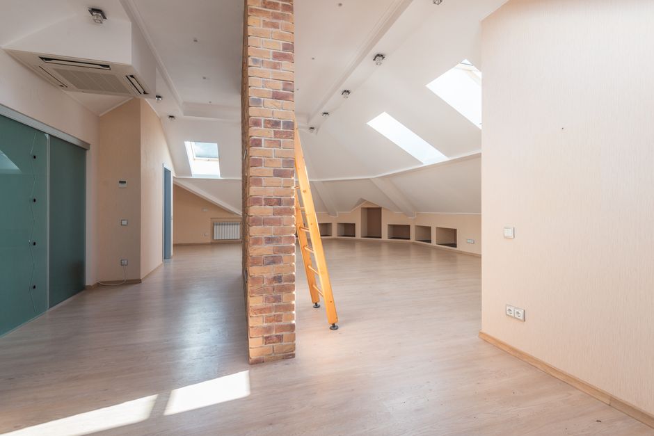 The image shows an expansive, modern attic or loft space with a sloped ceiling fitted with multiple skylight windows that allow natural light to brighten the area. The space features light-colored wooden flooring and pale cream or beige walls, creating a neutral, airy atmosphere. A prominent brick column runs vertically through the centre, partially dividing the room, with a yellow extension ladder leaning against it. To the left, there is a large, sliding, metallic storage cabinet with a greenish hue, and a visible electrical socket on the wall nearby. In the background, a small radiator is mounted along the sloped wall, and built-in shelving or storage compartments are set into the curved wall on the right side. The ceiling is fitted with a series of small, recessed spotlights, and an air conditioning unit is installed near the ceiling on the left. The space appears to be vacant, clean, and suitable for conversion or decoration, with clear indications that it is undergoing renovation or preparation for use, typical of a property in the vicinity of Bromley or similar South London suburbs. Rubbish Removal Bromley specializes in clearing such spaces, ensuring efficient removal of debris or renovation waste in the local area.