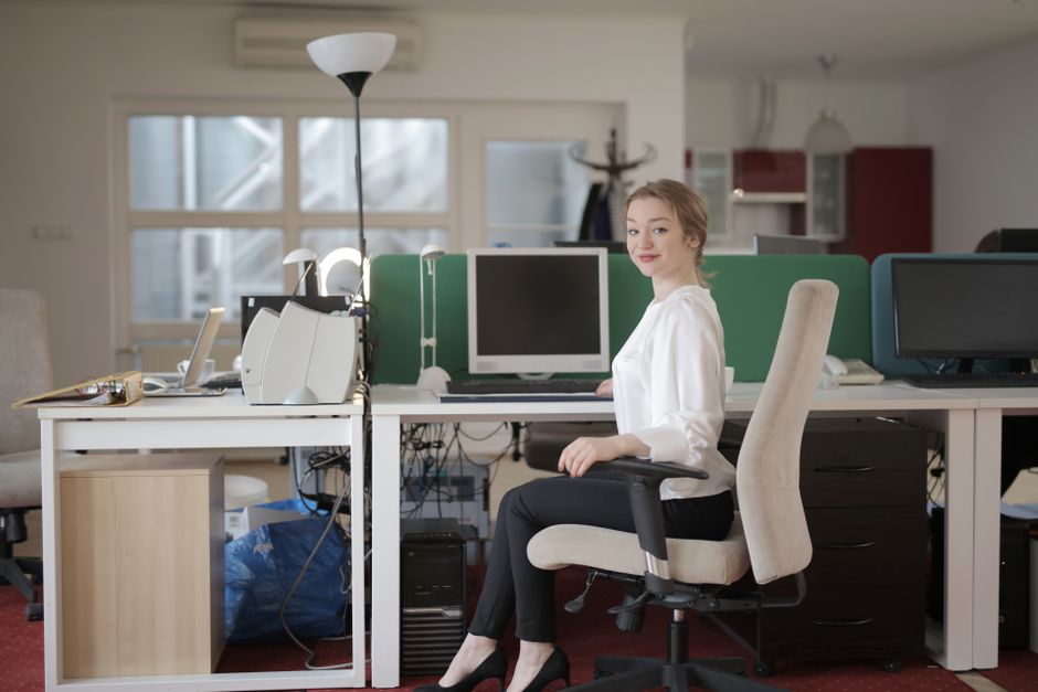In an office environment with a modern, open-plan layout, a young woman with blonde hair tied back, wearing a white blouse and black trousers, is seated in a beige ergonomic office chair facing a white desk. The desk is cluttered with various office equipment, including a white telephone, a laptop, and stacks of papers. Behind her, there are several computer monitors, some turned off, with green and black screens, and an adjustable desk lamp providing additional light. The background features a large window allowing natural daylight to illuminate the room, with a coat rack and kitchen area in the distance. The floor is covered with a red carpet, adding warmth to the professional setting, which is typical of contemporary office spaces in Bromley. The scene captures a moment of work at the desk, emphasizing a tidy but active workspace suited for administrative or clerical tasks, with no visible rubbish or waste in the scene. Rubbish Removal Bromley specialises in waste management services within this local area, although the image does not show any rubbish removal activity directly.