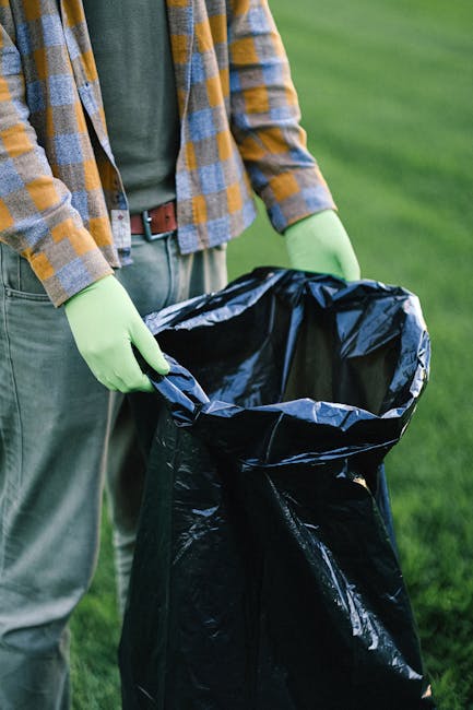 A person wearing a green long-sleeve shirt and a plaid jacket with shades of yellow, blue, and gray is holding open a large black rubbish bag lined with a silver inner lining, preparing for waste disposal. The individual is standing outdoors on a grassy area, on what appears to be a driveway or pavement, with blurred green grass in the background. The person is also wearing green gloves, indicating they are engaged in waste collection or rubbish removal activity. The scene suggests a tidy and organized approach to rubbish clearance, which could be related to garden waste or household waste management in Bromley. The wooden clothing and the environment reflect a professional yet casual setting typical of rubbish collection services. Rubbish Removal Bromley often handles such outdoor waste disposal tasks efficiently across Bromley, providing reliable rubbish clearance services in the area.