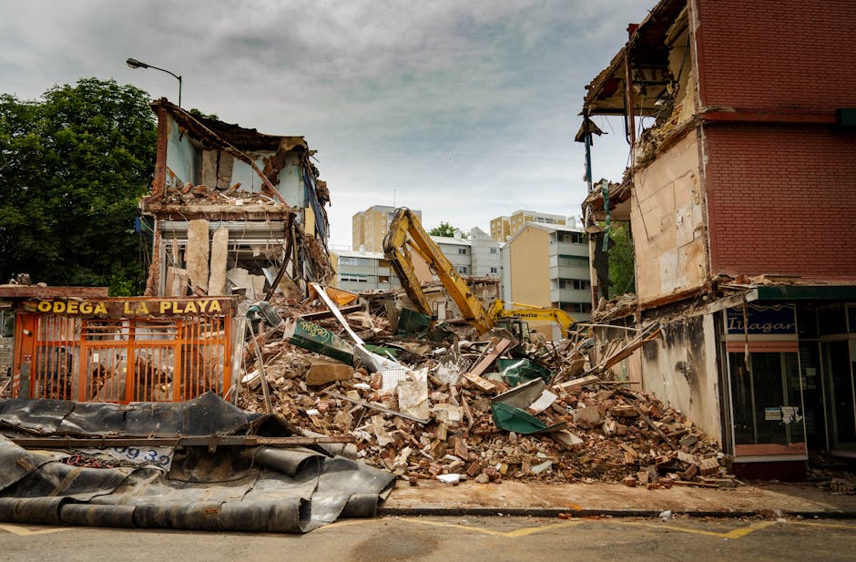 A large pile of debris and rubble from partially demolished buildings occupies the centre of the image, consisting of broken bricks, concrete fragments, wooden beams, and assorted construction materials. The rubble is spread across a paved area, with some materials stacked or leaning against remaining structures on either side. On the left, there is an orange metal barrier with the words 'Odega La Playa' printed in yellow, partially covered by debris. A yellow excavator arm is visible in the background, actively working amidst the destruction, with its hydraulic arm partially extended and digging into the debris. To the right, part of a red-brick building with signs of fire damage or scaffolding is visible, showing exposed and charred section walls. Behind the site, multi-storey apartment buildings with light-colored facades can be seen, indicating an urban environment. The sky above is overcast with a mix of grey clouds, creating a muted and neutral lighting atmosphere. This scene reflects a demolition or clearance process in a residential or commercial area, consistent with rubbish removal services in Bromley or nearby areas.